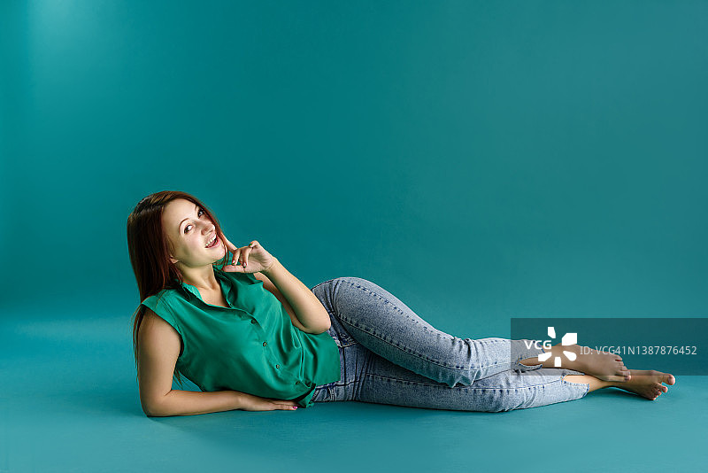 可爱的肖像漂亮迷人的年轻女子在绿色的衬衫和牛仔裤躺在蓝色的背景。图片素材
