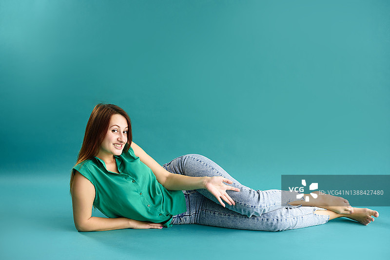 可爱的肖像漂亮迷人的年轻女子在绿色的衬衫和牛仔裤躺在蓝色的背景。图片素材