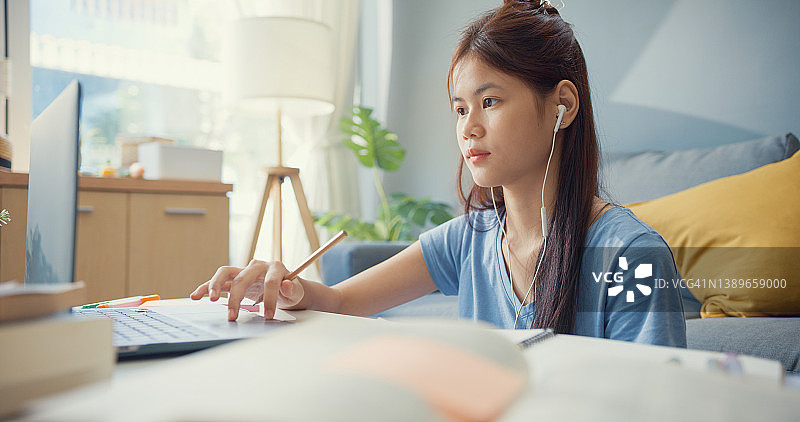 年轻的亚洲女孩十几岁，戴着休闲装耳机，用笔记本电脑上网学习，在客厅写讲稿。隔离教育在线电子学习冠状病毒大流行概念。图片素材