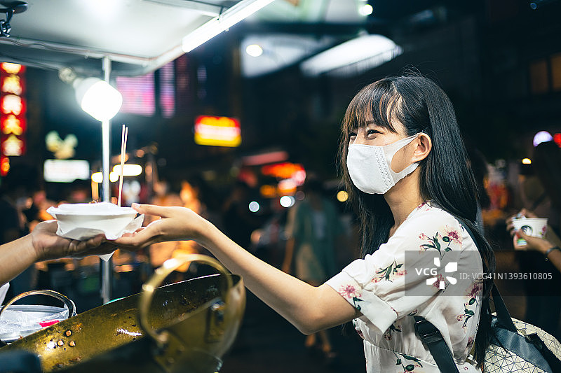 年轻的亚洲女性戴着口罩在曼谷夜市购买街头小吃图片素材