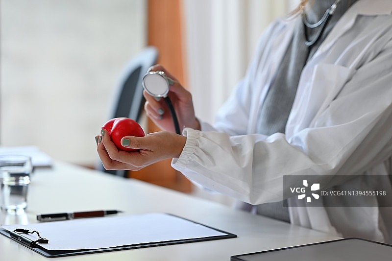 一个穿着白大褂的剖面图医生坐在办公室里，拿着一个心脏模型，用听诊器做一个检查姿势。为医疗保健理念。图片素材