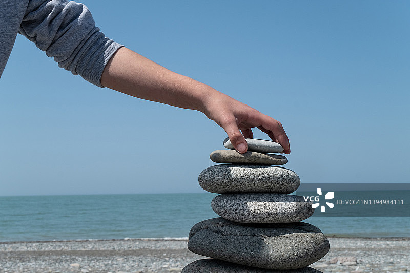 一个男人或少年在海滩上，背对着地平线，用海石或大卵石制作、建造和创造一个金字塔。灵魂中和谐与平衡的概念。一个人手里拿着一块石头，试图找到平衡。图片素材