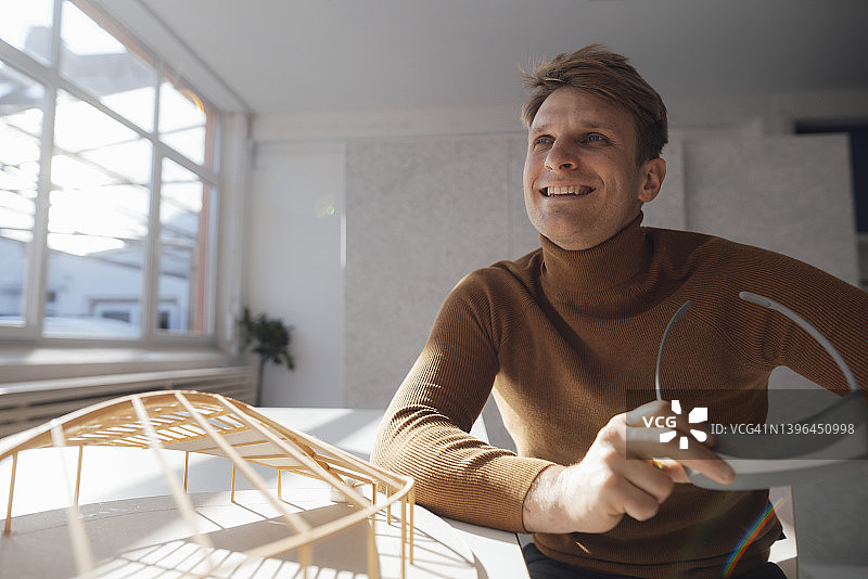 微笑的建筑师拿着虚拟现实模拟器坐在办公室的桌子形状的模型图片素材