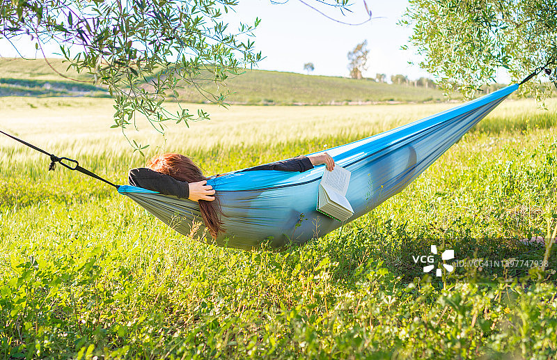 在夏天阳光明媚的田野里，匿名轻松女性睡在吊床上与书的侧面图片素材