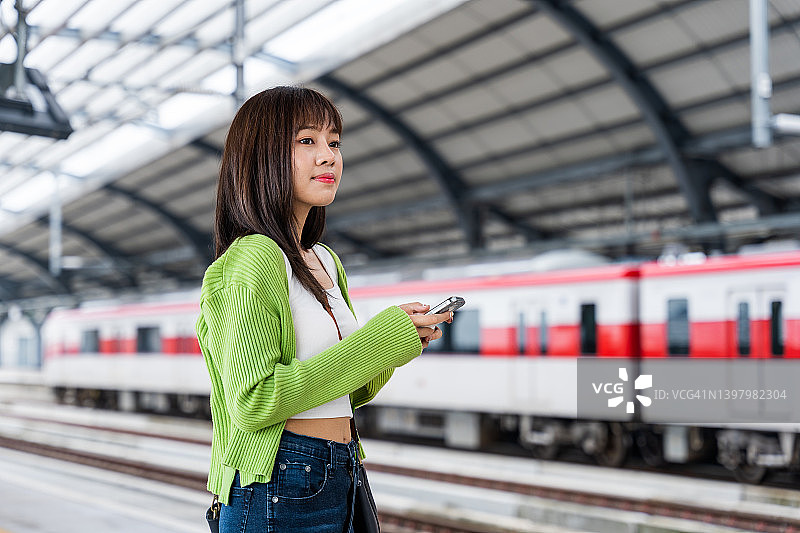一名年轻的亚洲女子在站台上等车时正在看智能手机。图片素材