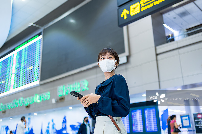 在机场候机楼，一名亚洲年轻女子在飞机时刻表前使用智能手机图片素材