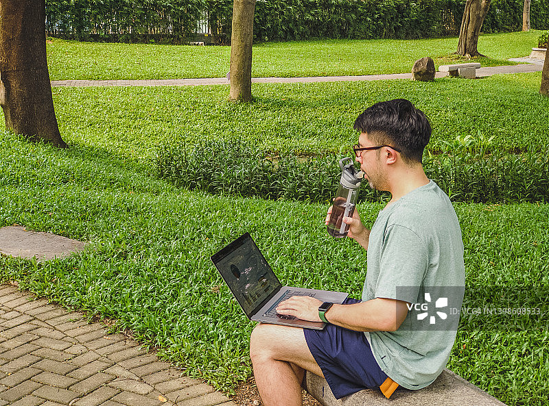 亚洲男子穿着运动服坐在长椅上使用笔记本电脑，在公园里用水瓶喝水-(在公园运动后工作)图片素材