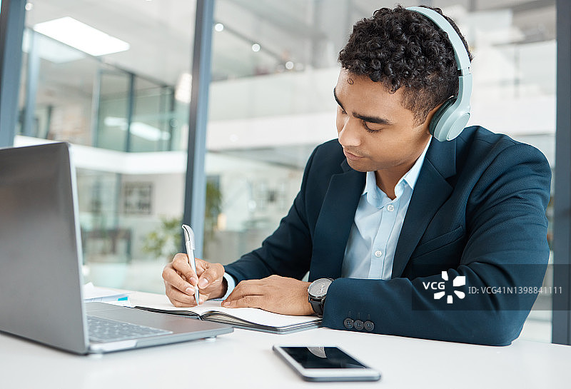 年轻严肃的混血商人，工作时戴着耳机，一边听音乐，一边在笔记本上写想法。一位西班牙裔男性商人在办公室里写日记图片素材