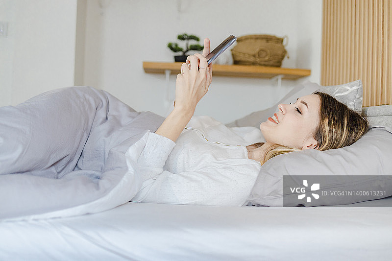 一个微笑着的年轻女人穿着睡衣躺在床上，她醒来后正在使用手机图片素材