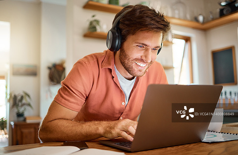 年轻开朗的白人商人戴着耳机，一边听音乐，一边独自在家使用笔记本电脑。一名男性商务人士在家厨房工作时在笔记本电脑上打字图片素材