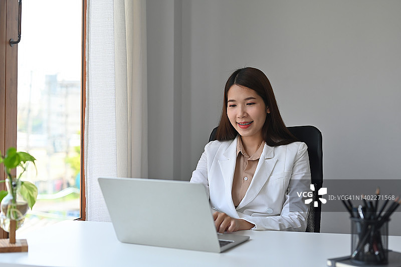 一个微笑的漂亮的亚洲商人坐在办公室，在笔记本电脑上工作的肖像，为商业和技术概念。图片素材