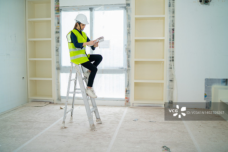 作为工程师或经理的亚裔女性在建筑工地有白墙和玻璃窗的房间里绘制房屋平面图，坐在铝梯上工作图片素材