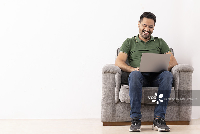 一个快乐的年轻人坐在沙发上使用笔记本电脑的肖像图片素材