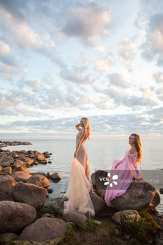 两个美丽的年轻华丽的女人的肖像穿着时尚的丝绸衣服对石头，日落的天空和云图片素材