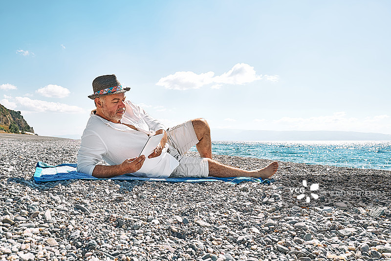 蓄着胡须的中年男子在沙滩上休息，用平板电脑阅读电子书。暑假读书和放松的概念。海滩上的文学图片素材