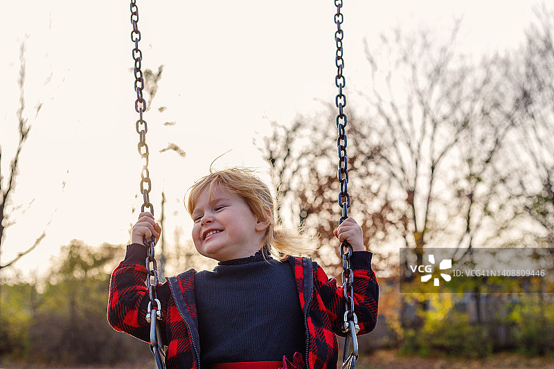 秋天，快乐微笑的小男孩坐在公园的秋千上图片素材