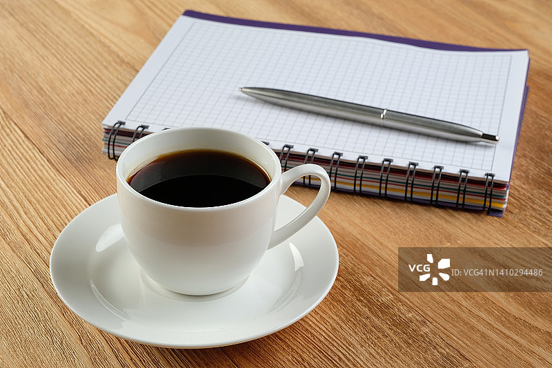 一支笔在一张空白的记事本上，一杯黑咖啡在一张木制的餐桌上。一个商人或经理在工作场所或在桌面上的项目。咖啡休息时间或商务会议，制定一天的计划。企业管理与发展。图片素材