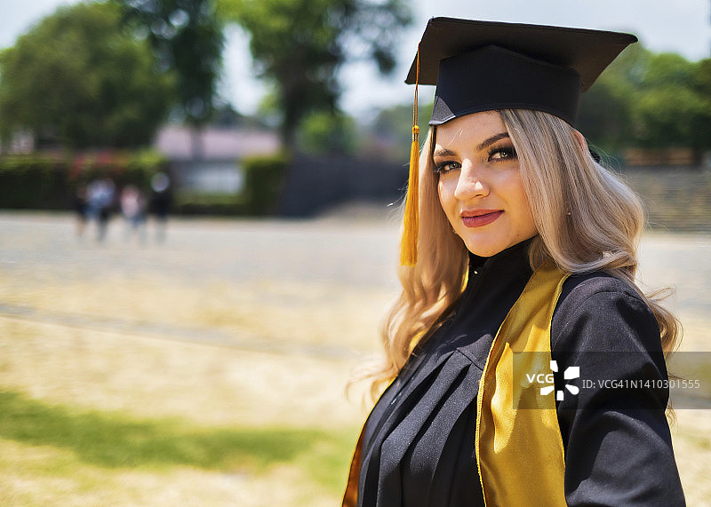 年轻的金发拉丁裔女子从大学毕业，微笑着看着镜头戴着毕业帽和毕业礼服图片素材