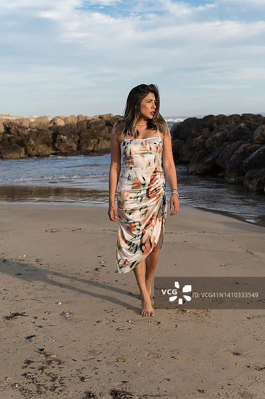 穿着裙子的年轻女子走在沙滩上，侧身看着。图片素材