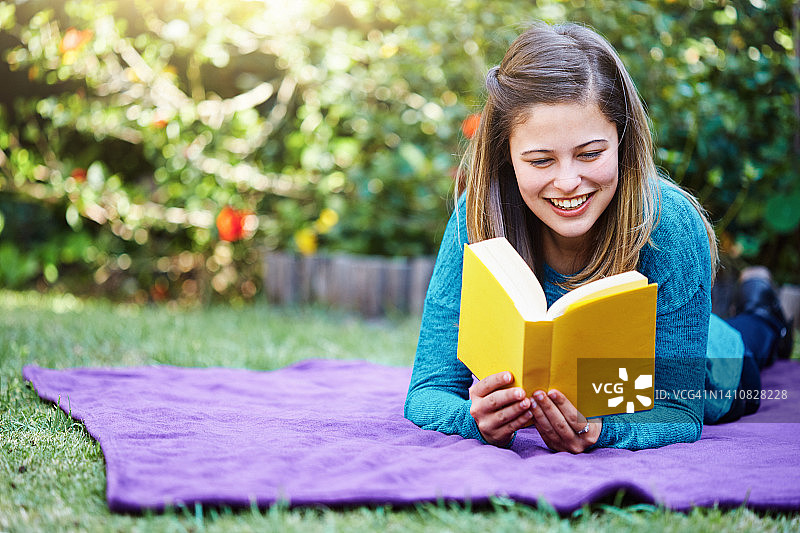 美丽的年轻女子躺在花园里的毯子上看书，看起来很开心很放松图片素材