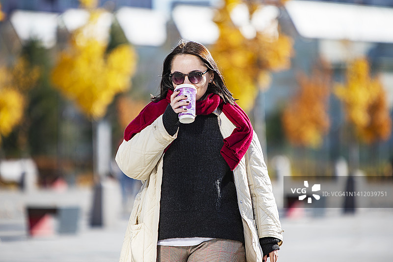 一个年轻的成年女子拿着咖啡纸杯走在秋天的公园里图片素材