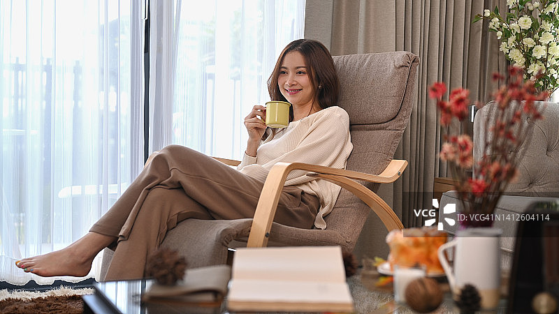 漂亮的年轻女子享受她的早晨咖啡，同时放松在舒适的扶手椅在家里。休闲活动，积极情绪概念。图片素材