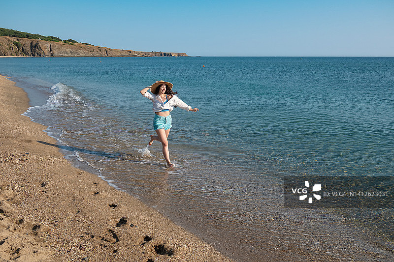戴着草帽的年轻快乐的女人在海滩上奔跑，在浅水中嬉戏图片素材