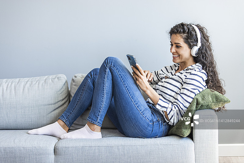 女人戴着耳机，拿着智能手机，坐在家里的沙发上做手势。图片素材