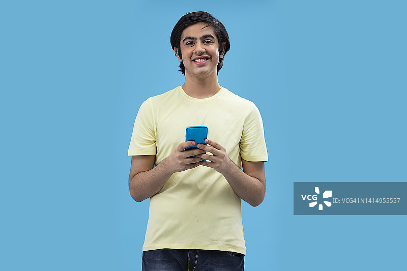 一个快乐的少年肖像使用智能手机，站在蓝色的背景图片素材