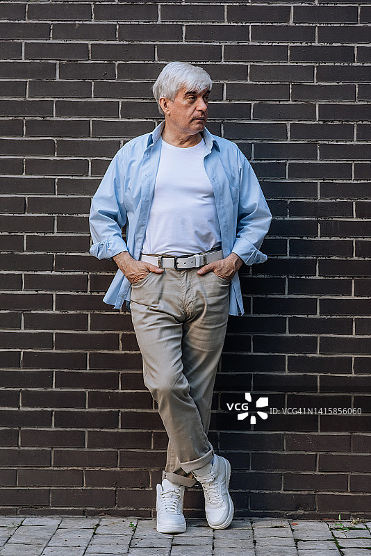 一个白发苍苍的老男人的全长垂直肖像摆姿势靠在一面墙在便装图片素材