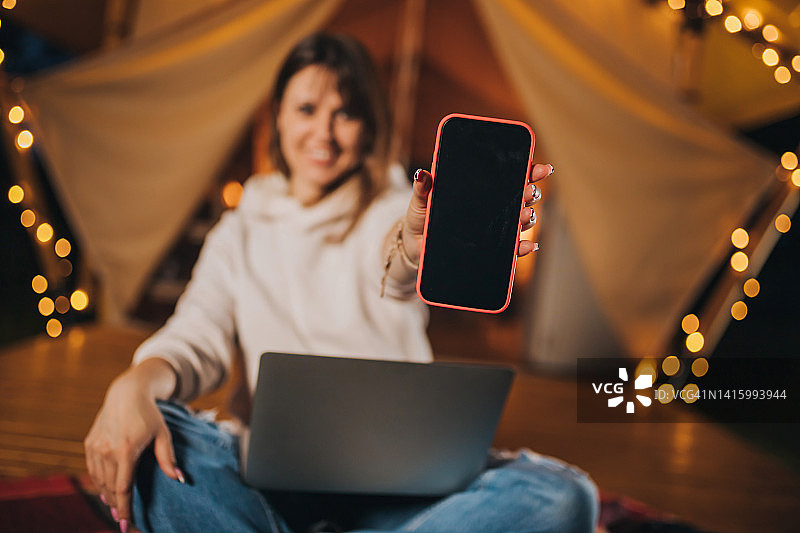 快乐女人自由职业者展示手机，同时工作的笔记本电脑在舒适的露营帐篷在夏夜。用于户外度假和度假的豪华露营帐篷。生活方式的概念图片素材