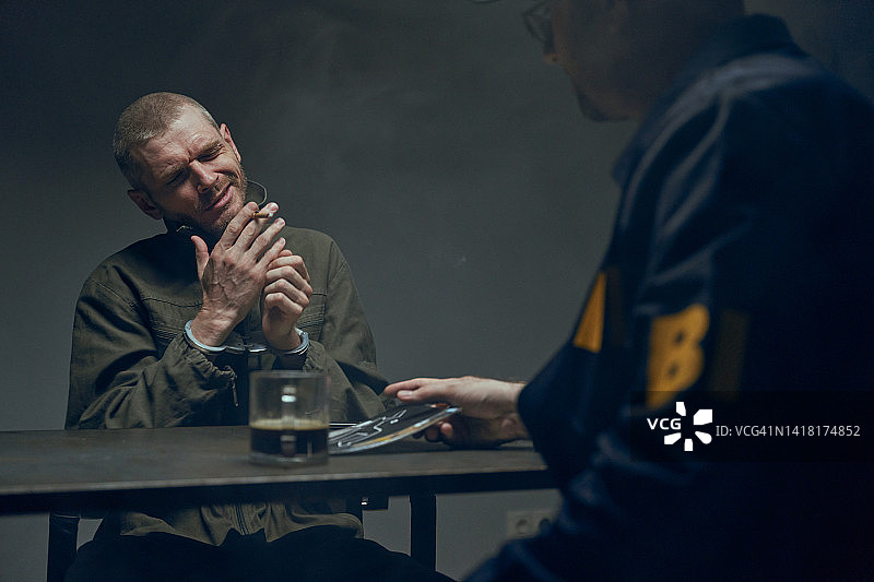 在一间黑暗的房间里，一个拿着香烟的小偷坐在警官面前的桌子旁图片素材