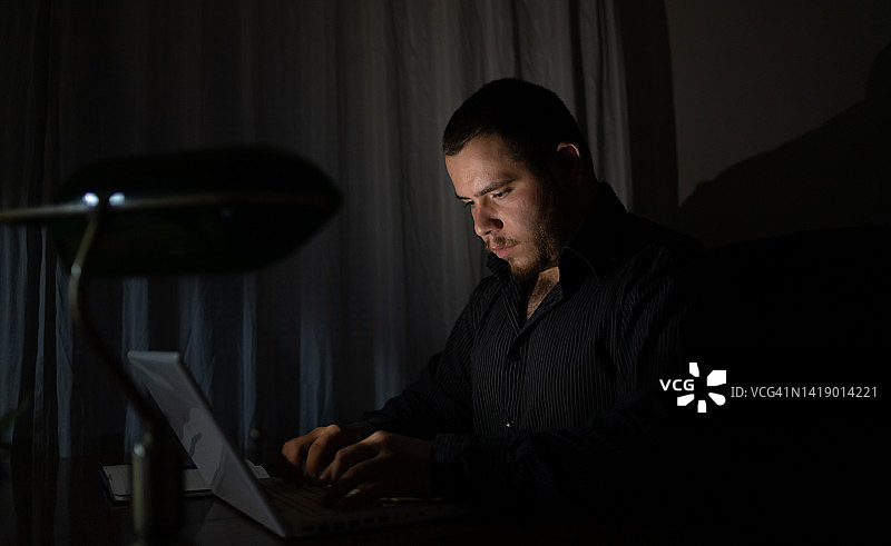 一个年轻人晚上在笔记本电脑前完成他的工作。图片素材