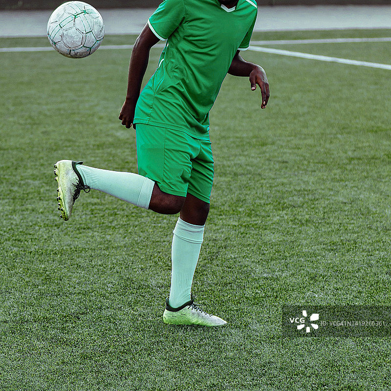 一个难以辨认的深色皮肤的人在绿色足球运动制服举起一个足球，同时在足球场上做熟练的技巧的特写图片素材