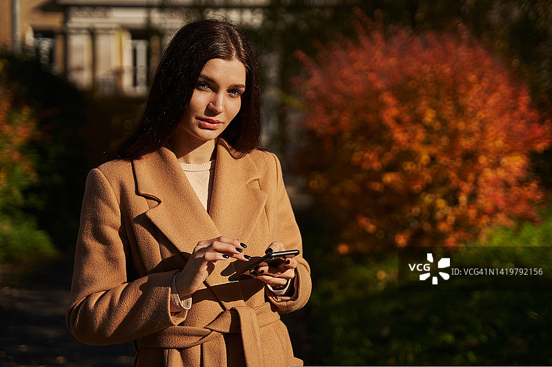 秋天的公园里，在美丽的红色灌木旁，一位穿着浅色大衣的美丽女子在阳光下看着智能手机里的东西图片素材