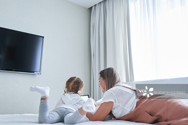妈妈和小女儿躺在靠窗的一张白色床上，对面是白墙，旁边放着电视和电子平板电脑图片素材