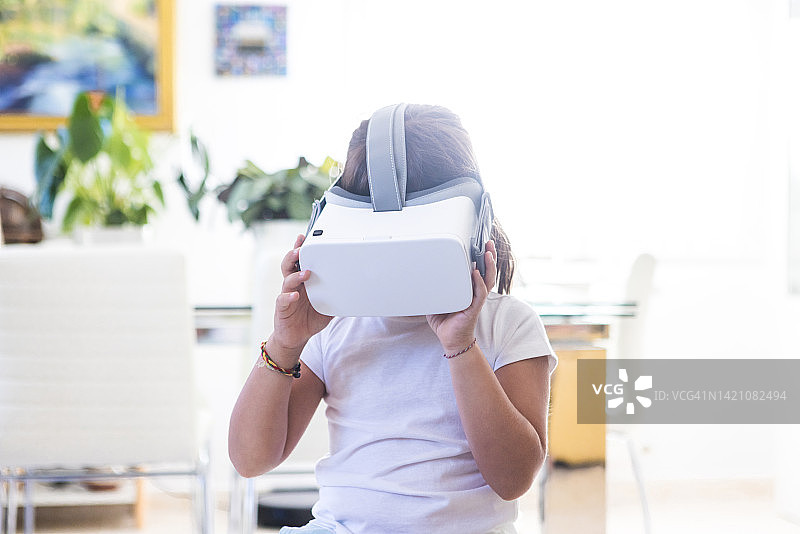 女孩在她的客厅里戴着虚拟现实眼镜，低头看视频，用她的手拿着眼镜。概念虚拟现实，互动，虚拟模拟器，技术和未来。图片素材
