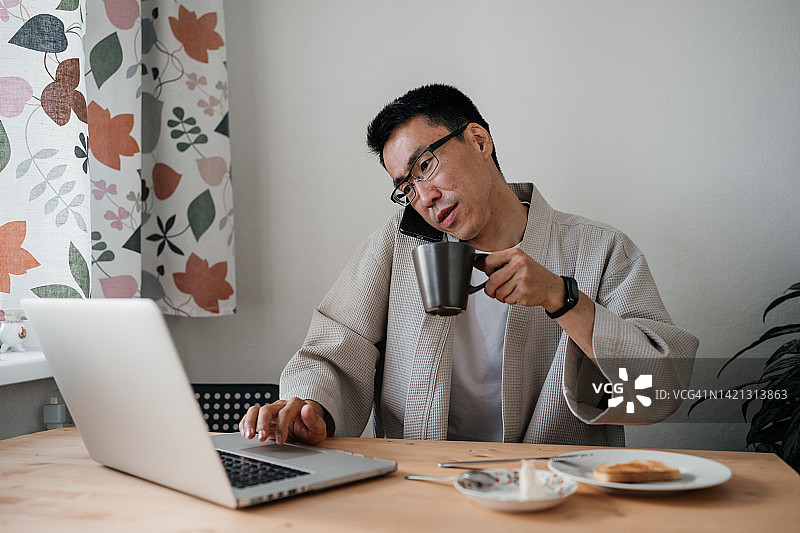 亚洲中年男子戴眼镜，在厨房里用智能手机和笔记本电脑。图片素材