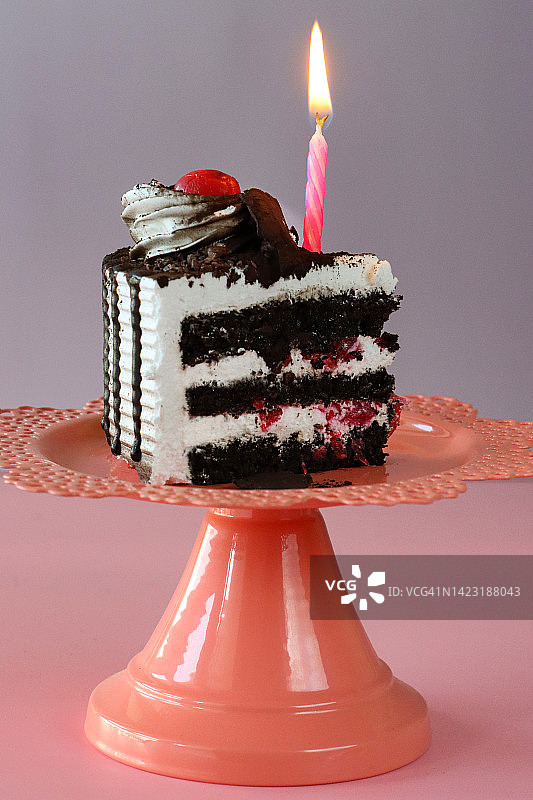 黑森林蛋糕片的图像，三层蛋糕层被橙色蛋糕架上的鲜奶油隔开，点燃的生日蜡烛，管状鲜奶油花环，莫雷罗樱桃，豪华巧克力蛋糕，巧克力刨花，粉紫色的背景，重点在前景图片素材