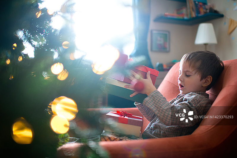 可爱的小男孩穿着睡衣，拿着圣诞礼物，坐在家里的圣诞树旁图片素材