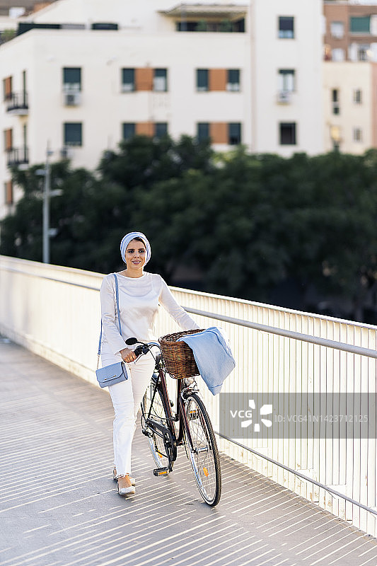 一位穆斯林妇女推着自行车走在人行道上，望向别处图片素材
