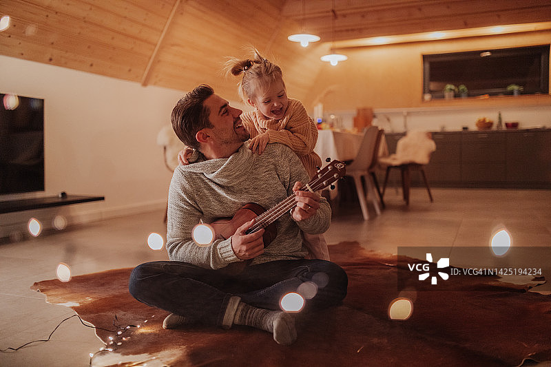 年轻快乐的爸爸和他的女儿坐在客厅里弹吉他。图片素材