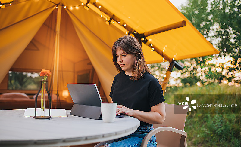 快乐的女性自由职业者在一个阳光灿烂的日子里在舒适的野营帐篷上使用笔记本电脑。豪华户外露营帐篷，适合夏季度假和度假。生活方式的概念图片素材
