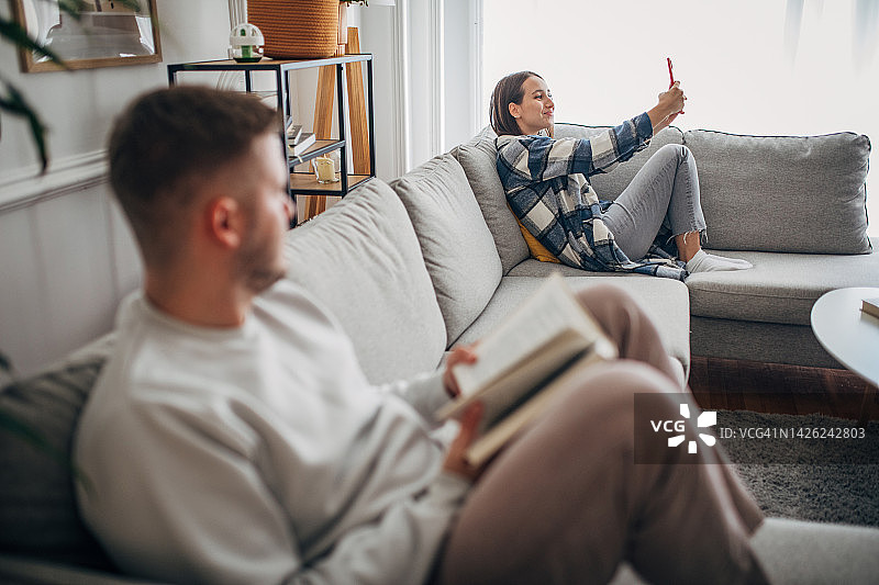 年轻男子在家里的沙发上看书，而他的女朋友正在用智能手机上网图片素材