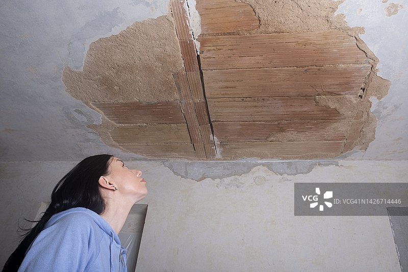 一名年轻女子抬头看着天花板上被毁的洞。灰泥因受潮而脱落。旧公寓的修复和修理。图片素材