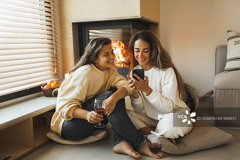 两个年轻的女性朋友在壁炉边放松，笑着，喝着酒，用手机刷着社交媒体图片素材
