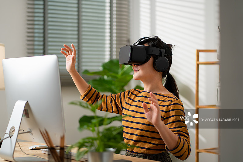 亚洲女性戴着VR头戴设备，在空中触摸，在家里办公室微笑。图片素材