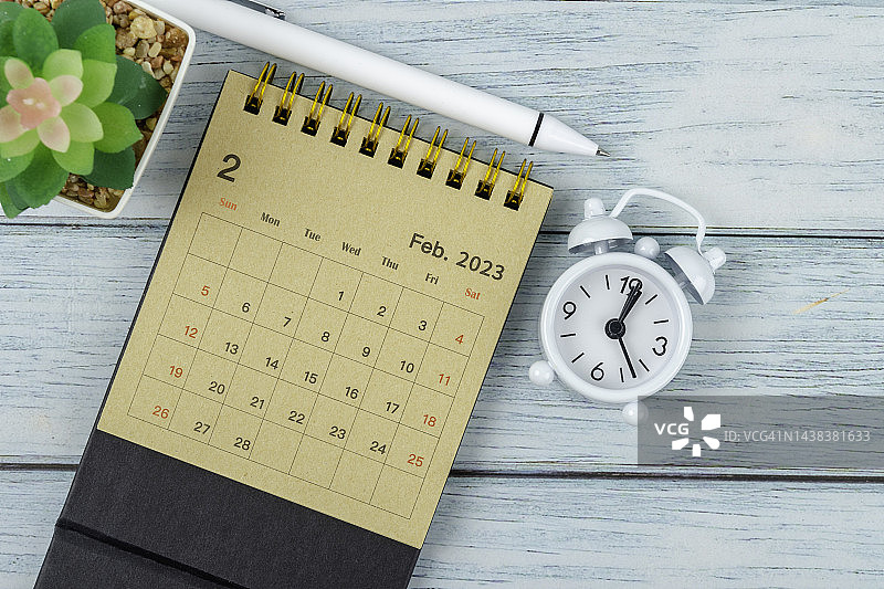 日历桌2023:2月是组织者计划和截止日期的月份，木桌背景上有一个闹钟和白笔。图片素材