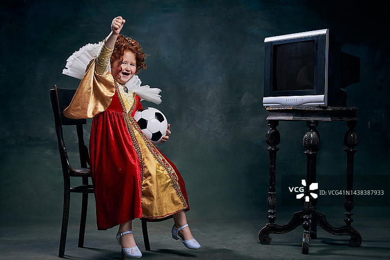 小红发女孩的肖像，穿着皇室人物服装的孩子在看足球比赛孤立在深绿色的背景图片素材