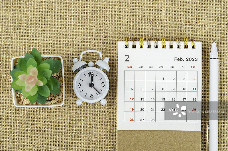 2023日历台:2月是组织者制定计划和期限的月份，有闹钟、室内植物和粗麻布背景的白色钢笔。图片素材